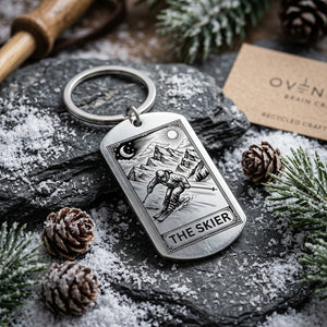 A silver metal keychain with a dog-tag-shaped pendant engraved with a skier and the words 'THE SKIER' and 'OVEN BRAIN', styled on a dark stone surface with pine needles, snow, pinecones, and a kraft paper tag.