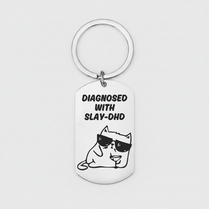 A silver metal dog tag keychain with a circular key ring, featuring a black graphic of a cartoon cat wearing sunglasses and the text 'DIAGNOSED WITH SLAY-DHD' on a light gray background.