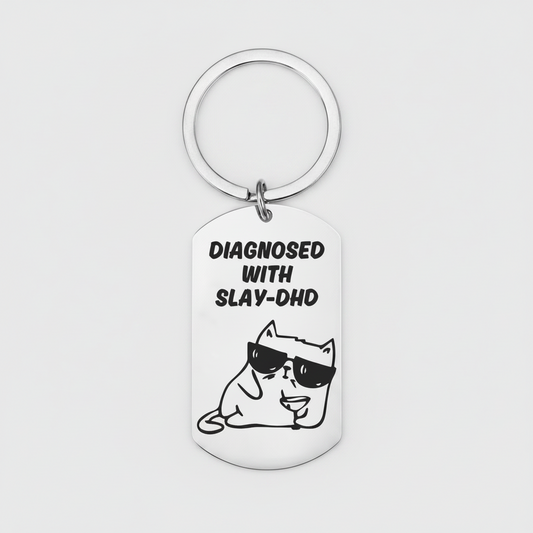 A silver metal dog tag keychain with a circular key ring, featuring a black graphic of a cartoon cat wearing sunglasses and the text 'DIAGNOSED WITH SLAY-DHD' on a light gray background.