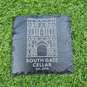 A square black slate coaster engraved with a white line drawing of South Gate Cellar architecture and the text 'SOUTH GATE CELLAR Est. 2019', placed on green grass.