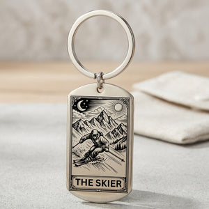 A silver-toned metal dog tag keychain with an engraved tarot-style illustration of a skier and mountains, labeled 'THE SKIER,' shown on a light surface with a soft blurred background.