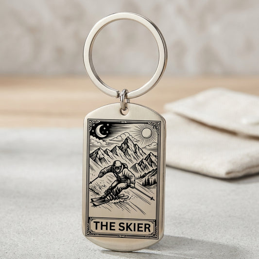 A silver-toned metal dog tag keychain with an engraved tarot-style illustration of a skier and mountains, labeled 'THE SKIER,' shown on a light surface with a soft blurred background.