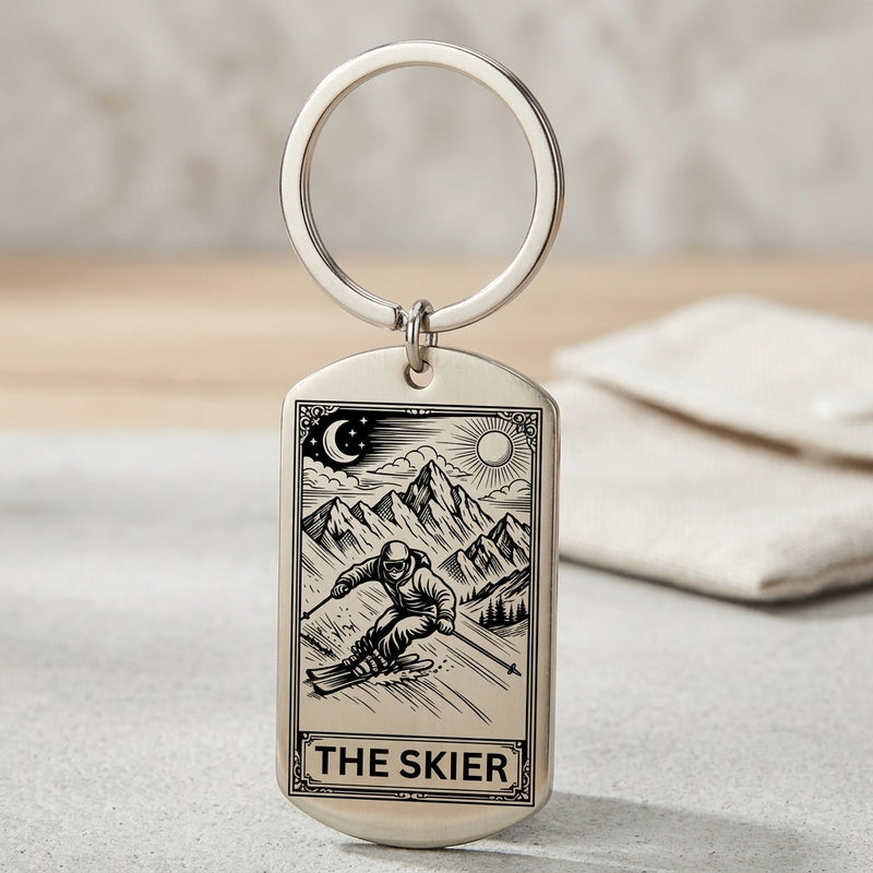 A silver-toned metal dog tag keychain with an engraved tarot-style illustration of a skier and mountains, labeled 'THE SKIER,' shown on a light surface with a soft blurred background.